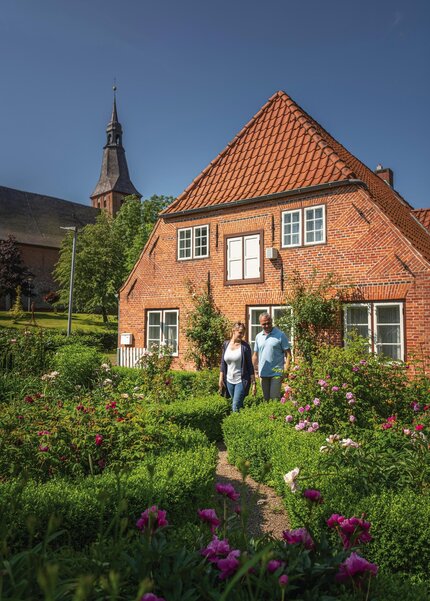 Paar spaziert durch grünen Garten vor imposantem roten Backsteinhaus