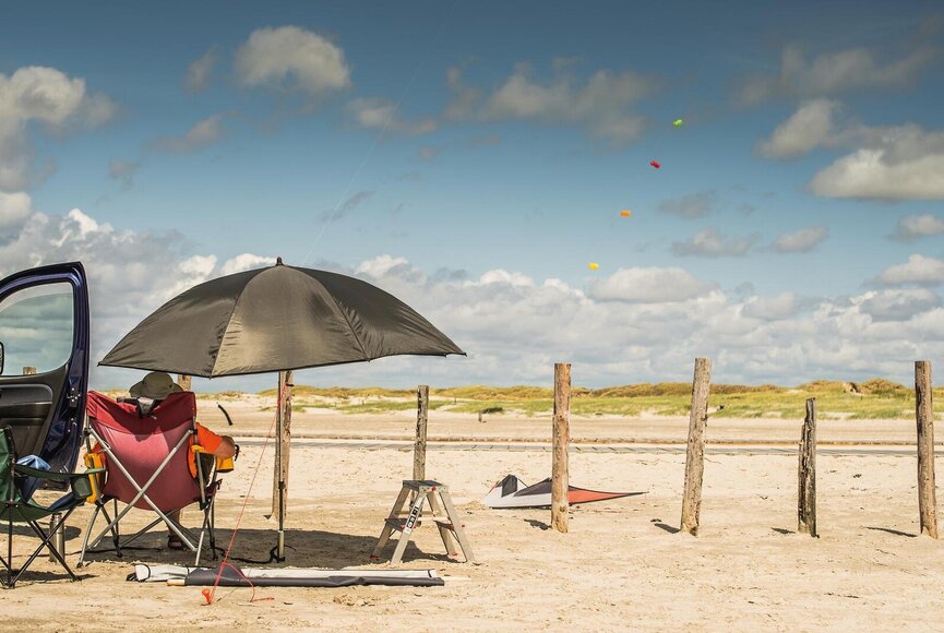 Person im Campingstuhl unterm Sonnenschirm am Strand mit Blick Richtung Strandparkplatz
