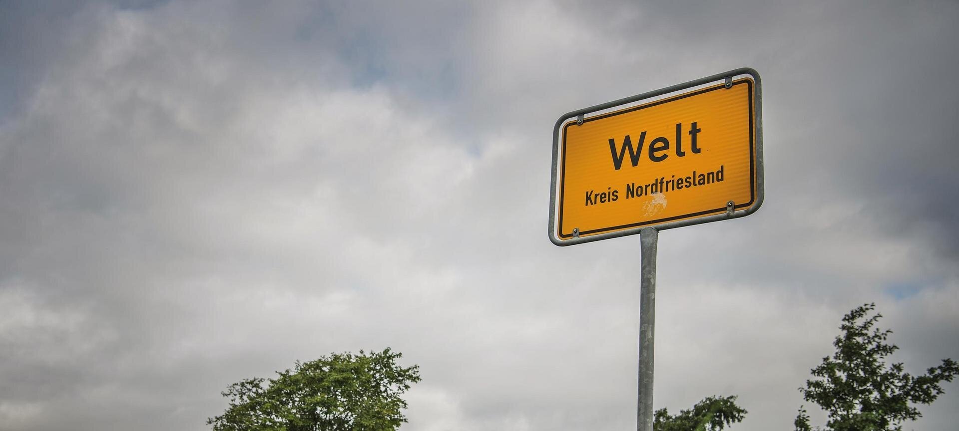 Ortseingangsschild von Welt mit Fahrradfahrern im Hintergrund