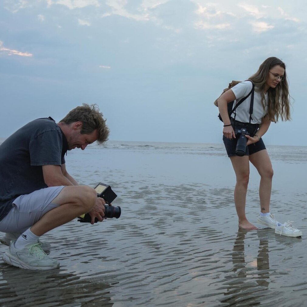 Marina zieht sich die Socken am Strand aus und wird dabei vom Kameramann gefilmt
