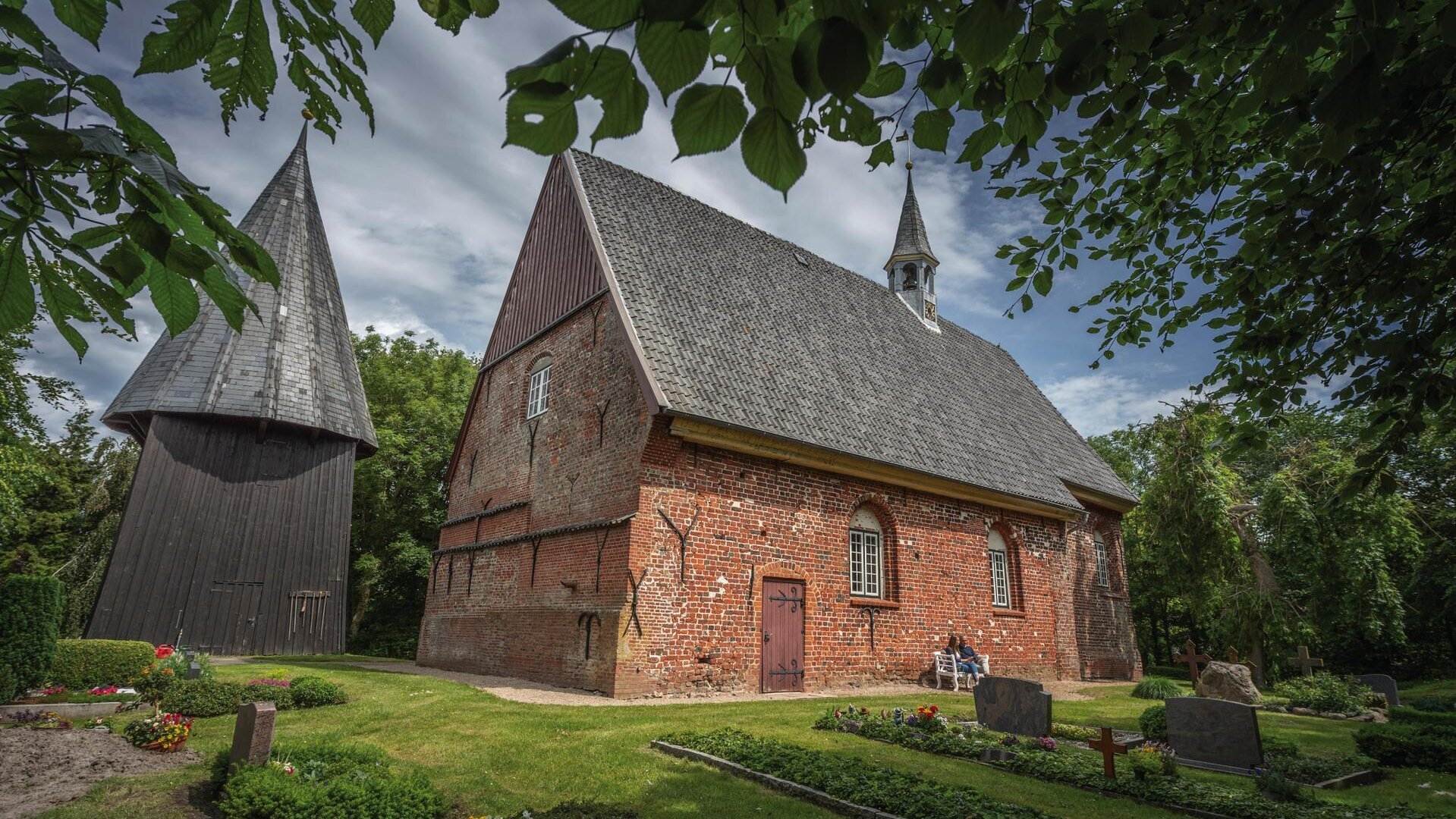 urige Kirche aus rotem Backstein mit dunklem Dach und kleiner Kirchturmspitze und separatem Turm daneben