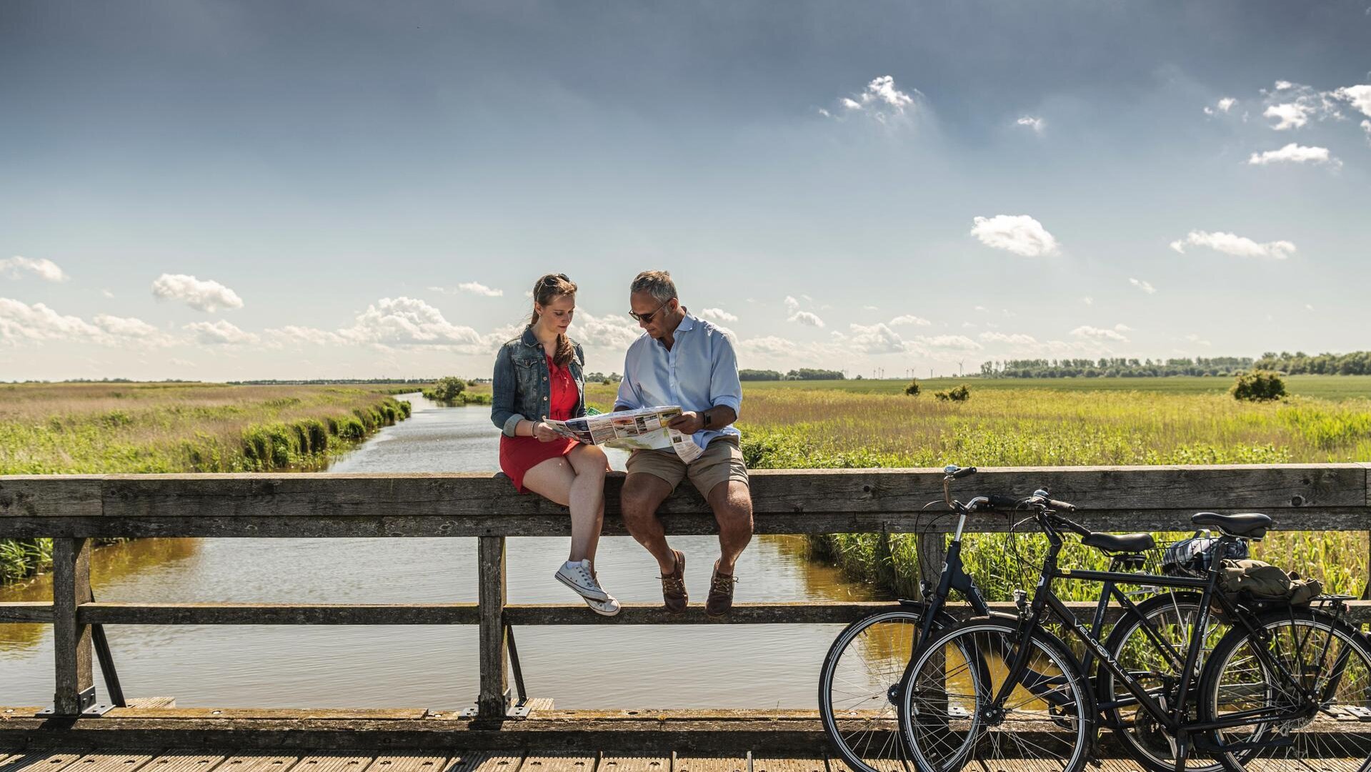 Paar sitzt bei Sonnenschein auf einem Geländer und schaut gemeinsam in eine Fahrradkarte