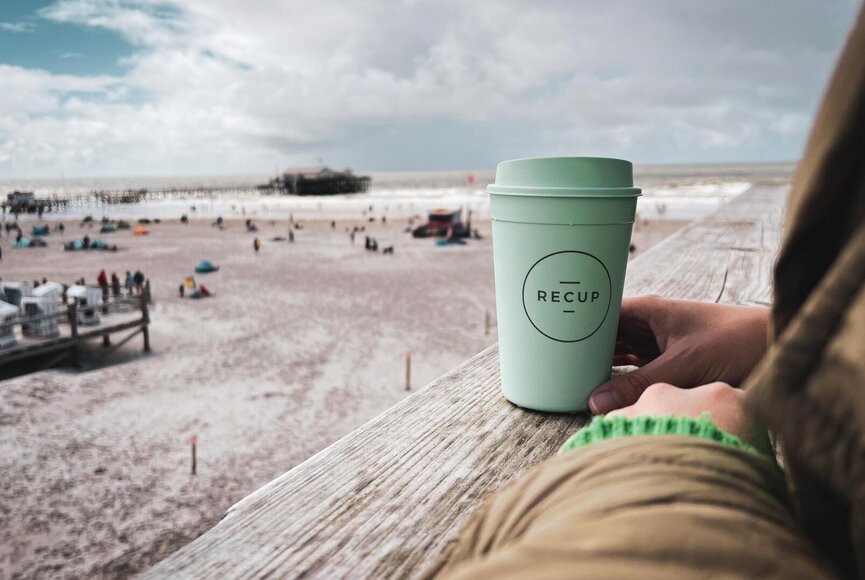 Frau mit RECUP-Becher auf einem Pfahlbau mit Blick auf den Strand