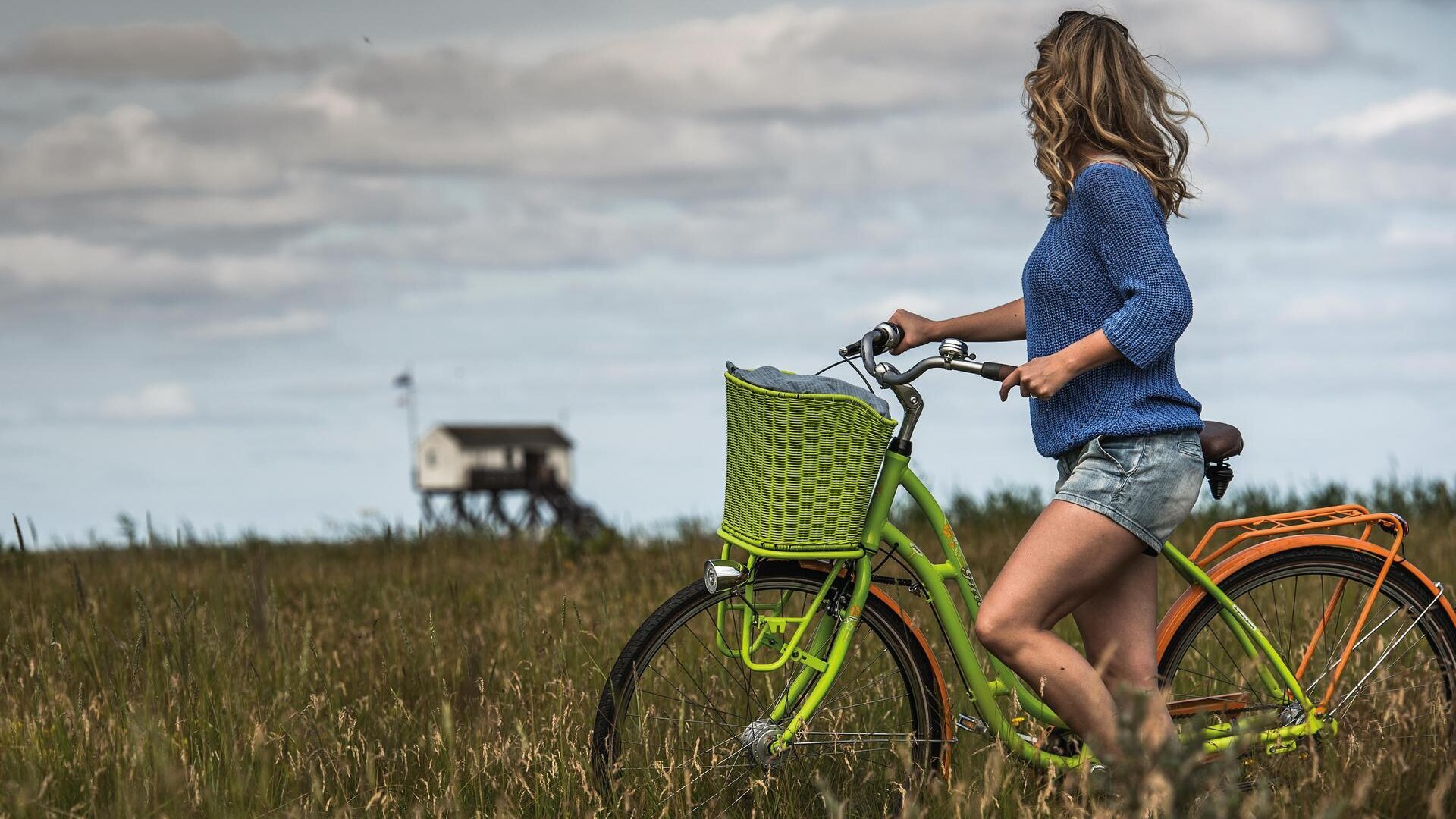 Frau mit grünem Fahrrad vor den Salzwiesen mit Pfahlbau im Hintergrund