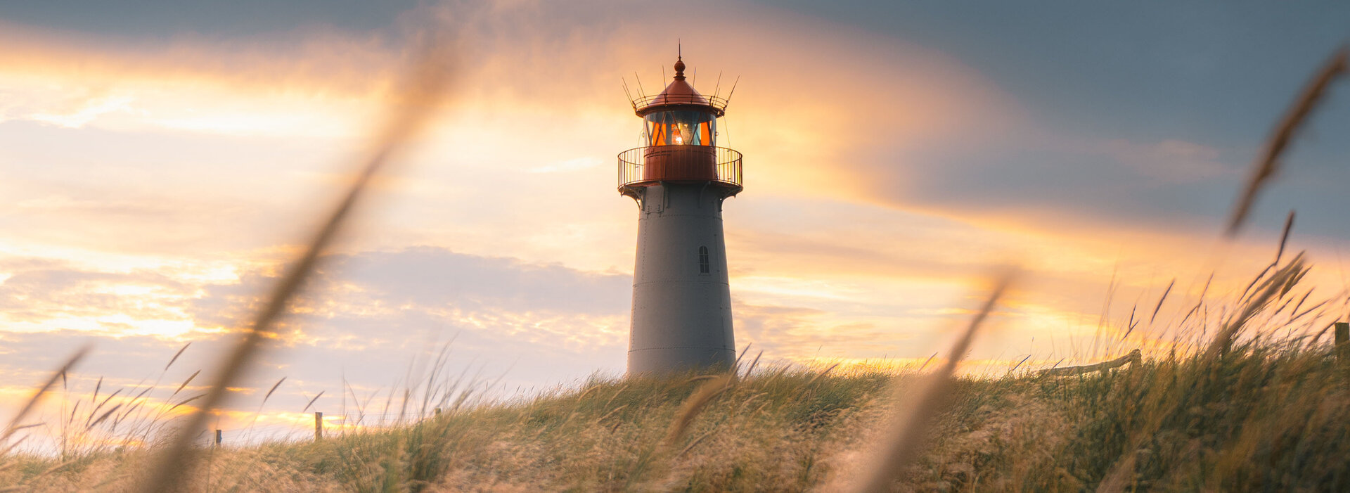 Der Sylter Leuchtturm List schaut hinterm Deich hervor. Im Vordergrund wehen Gräser.